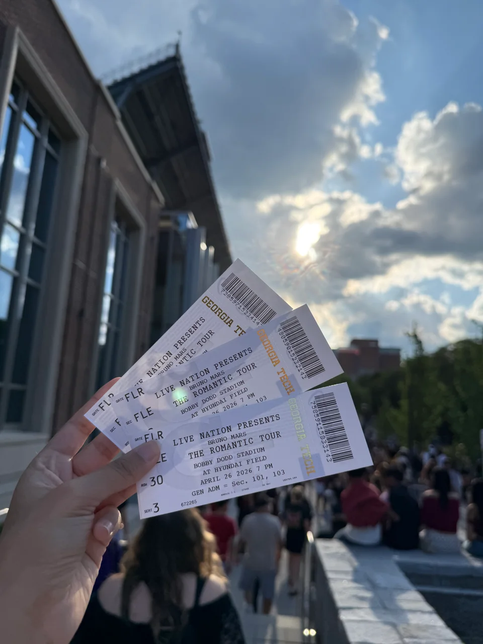 A hand fanning out three tickets to Bruno Mars’ Romantic Tour at Georgia Tech’s Bobby Dodd Stadium, under a sunny, cloudy sky.
