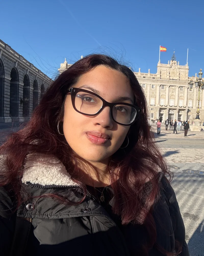 Anya in front of the Royal Palace of Madrid on a sunny day, the Spanish flag flying over the palace.