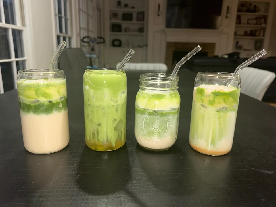 Four homemade iced matcha lattes in glass jars with glass straws, each layered a little differently.