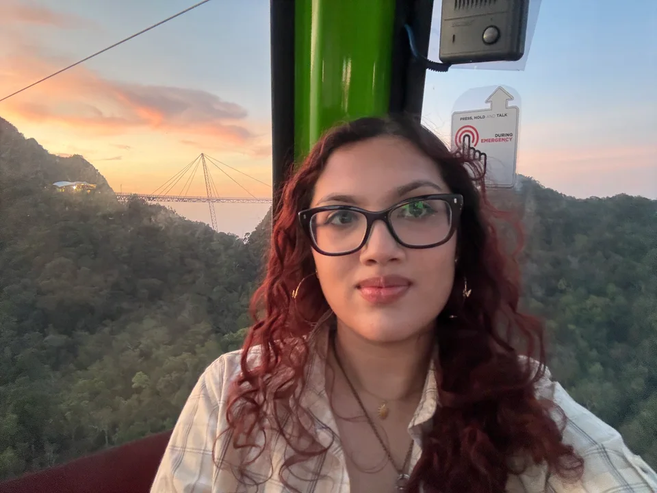 Anya in a cable car at sunset, with a curved suspension bridge over forested mountains behind her.