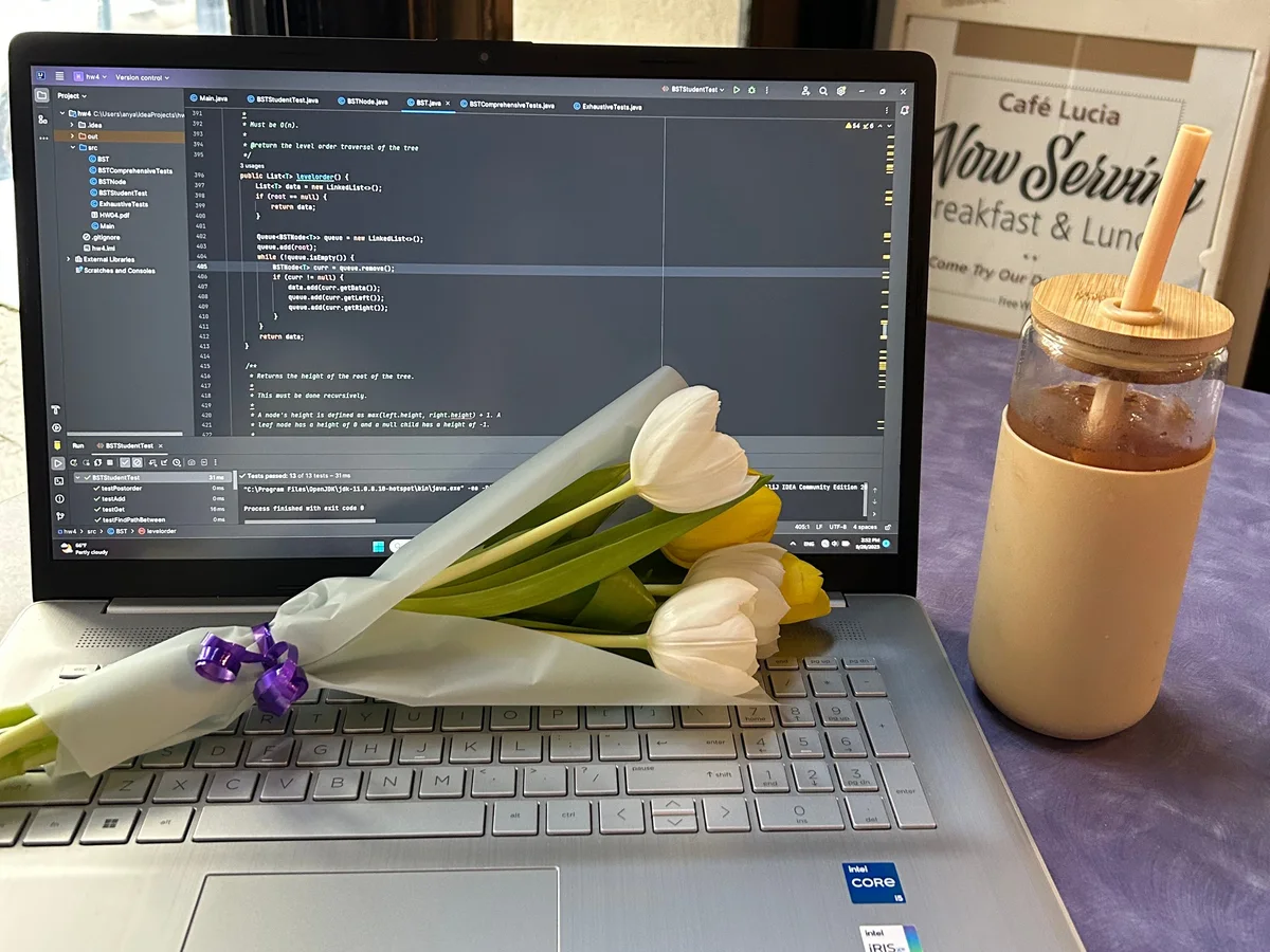 A laptop at a café showing Java code with all tests passing, a bouquet of white and yellow tulips across the keyboard and an iced drink beside it.
