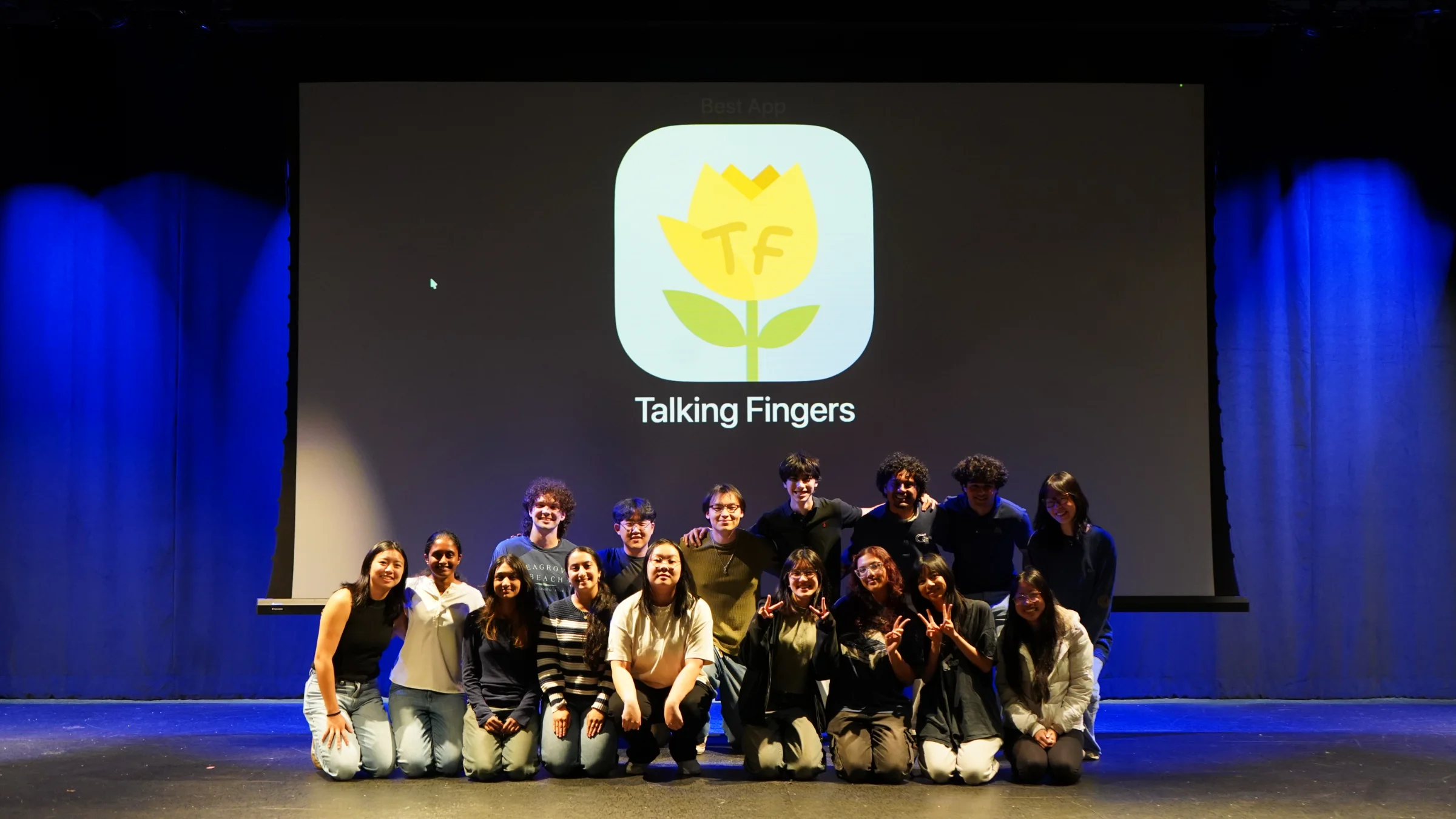 The Talking Fingers team on stage at Demo Day, in front of the app icon projected on a large screen.